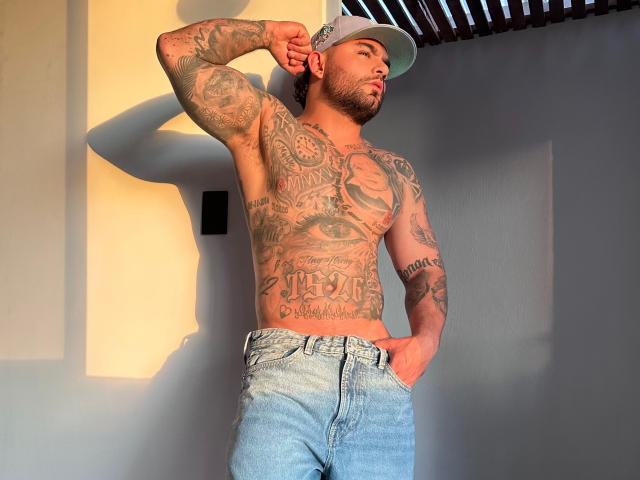 LyamVera sans chemise, avec des tatouages recouvrant sa poitrine et ses bras, portant une casquette et un jean bleu, une main dans la poche, debout au soleil, montrant un corps musclé, tatoué et barbu..