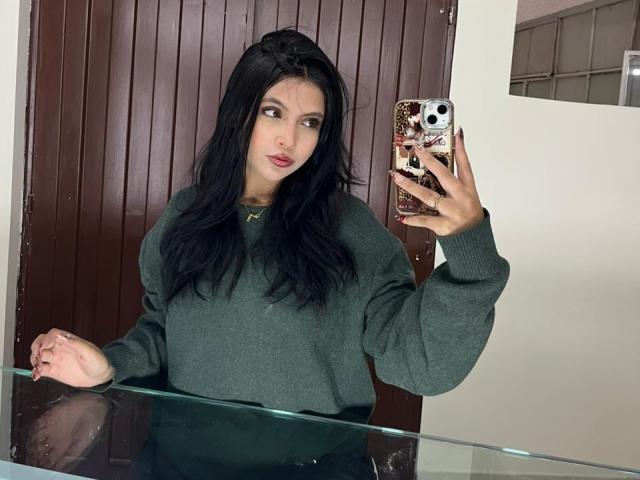 NyraVanel with long black hair, green sweater, holding phone with floral case, taking mirror selfie, looking to the side with a slight pout, white wall, wooden door.