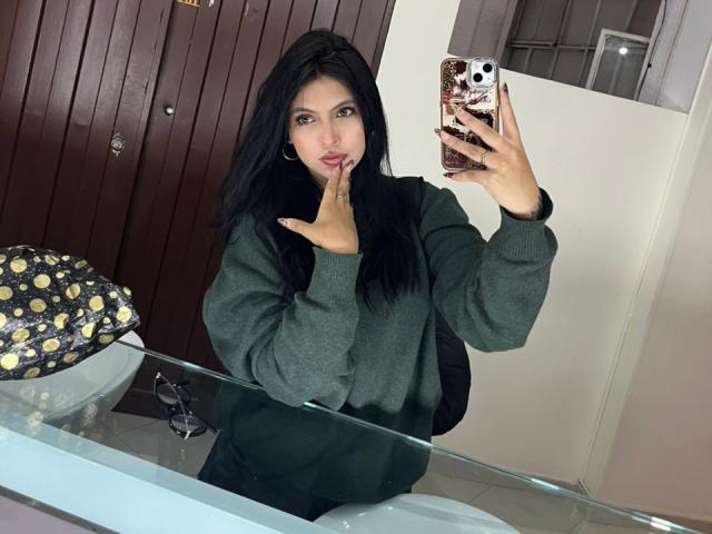 NyraVanel, con cabello negro largo, un suéter verde y pantalones negros, sosteniendo un teléfono, con el dedo en los labios, un selfie frente al espejo, una funda de teléfono con un diseño blanco y dorado, un fondo de puerta marrón..