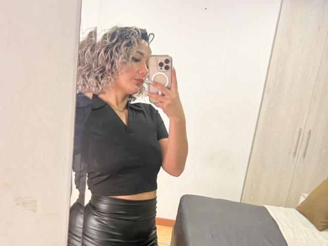 Flora Saenz mit lockigem, grauen Haar, einem schwarzen Crop-Top und einem schwarzen Lederrock, macht ein Selfie vor einem Spiegel mit ihrem Handy und zeigt dabei ihren Hintern und ihre kleinen Brüste..