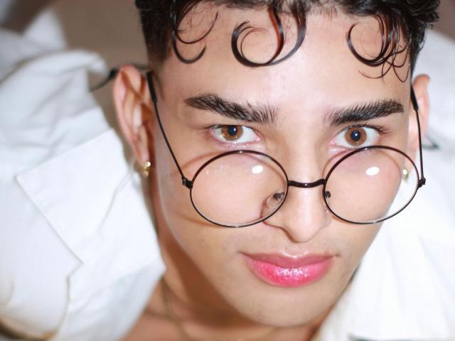 LogaanHotts, avec ses cheveux noirs bouclés, portant des lunettes rondes, a des lèvres roses brillantes et des yeux marron, il porte une chemise blanche et une boucle d'oreille dorée, et regarde légèrement vers le haut de l'appareil photo..
