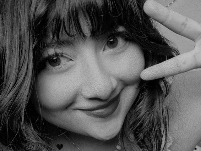 NekoPam ha i capelli scuri e ondulati, fa un gesto di pace con la mano destra, sorride mostrando leggermente i denti, in un selfie in bianco e nero. Il suo naso è piccolo e carino..