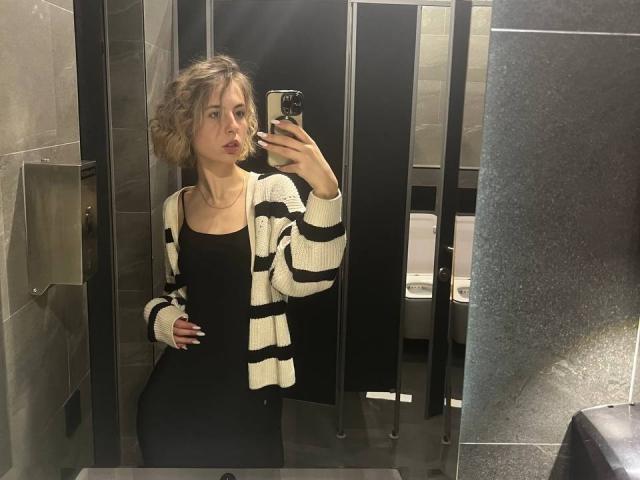 Tora Stambaugh, avec ses cheveux blonds bouclés, portant une robe noire et un cardigan à rayures blanches, prend un selfie devant un miroir dans une salle de bain, montrant sa petite poitrine et ses fesses discrètes..