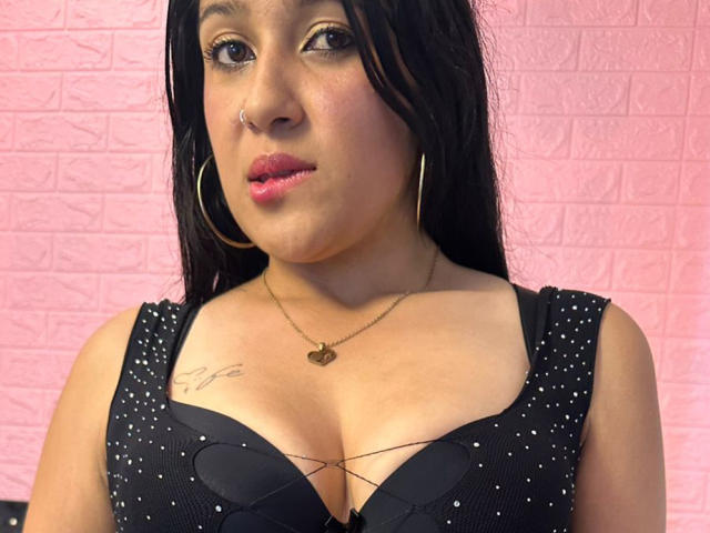 LuxyLeim heeft middelgrote borsten en draagt een zwarte, glinsterende, kruisende BH, gouden oorringen en een hartvormige ketting, met roze lippenstift en een neusring, en kijkt naar de camera..