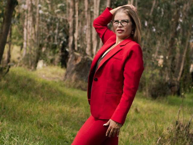 ParisMatur69, avec des cheveux blonds, des lunettes, une combinaison rouge, et une main dans les cheveux, debout dans une forêt verdoyante, montrant ses fesses, avec une expression confiante et un léger sourire..