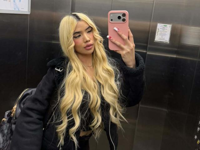 ChristieGarci with long blonde wavy hair, wearing a black fuzzy jacket, taking a selfie in a mirror, holding a pink phone, with a black bag on her shoulder.