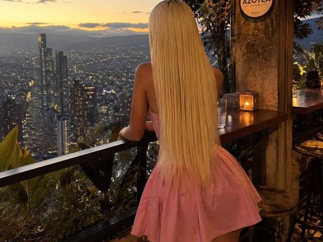 ChristieGarci, com longos cabelos loiros, vestindo um vestido rosa, está de costas para a câmera, olhando o horizonte da cidade a partir de um bar no terraço, com as nádegas levemente elevadas, e uma mesa com velas à direita..