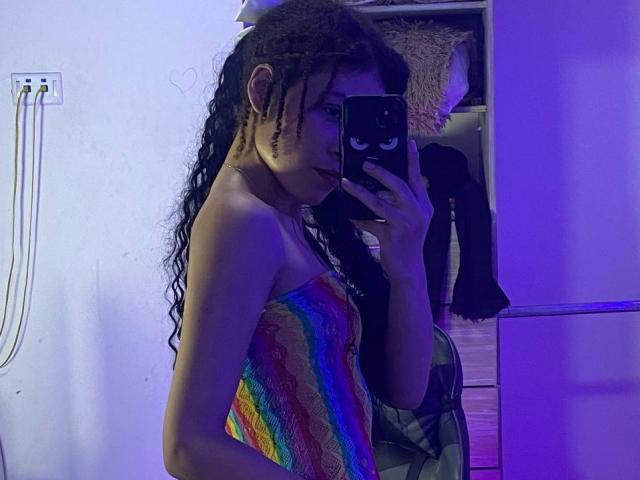 La tua Liliana, con lunghi capelli neri ricci, indossa un abito senza spalline a righe arcobaleno e sta scattando un selfie allo specchio con un telefono che ha un simpatico adesivo a forma di faccina. L'illuminazione della stanza è viola..