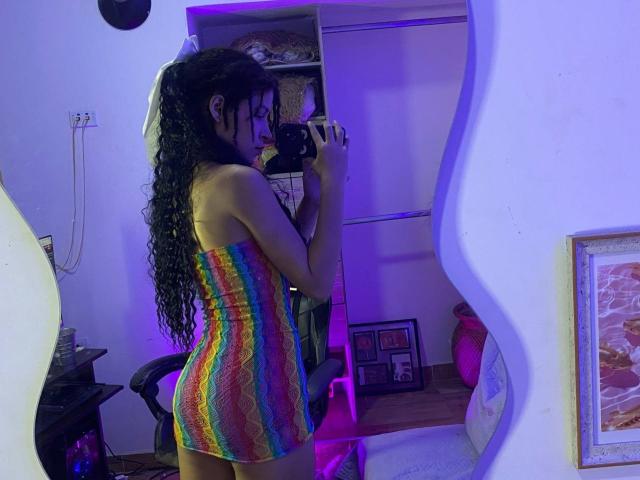 YourLiliana with curly black hair, wearing a rainbow-striped, strapless, tight mini-dress, holding a camera in a mirror selfie, showing her ass and small tits. Purple room lighting.