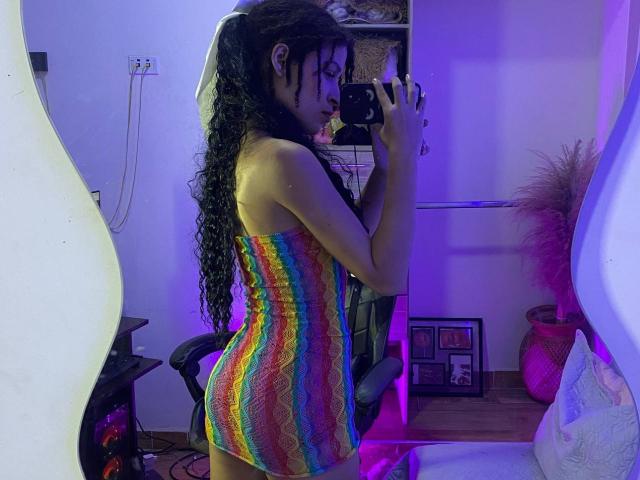 La tua Liliana, con lunghi capelli neri ricci, indossa un vestito aderente e strutturato a righe arcobaleno, tiene in mano un telefono, sta scattando un selfie allo specchio, mostrando il suo piccolo sedere sodo e il suo corpo slanciato e sensuale..