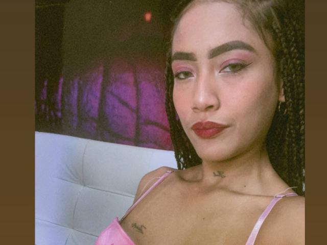SusyXpleasure has light brown skin, red lipstick, pink eyeshadow, and braided hair. She's wearing a pink top, showing small tits and a star tattoo on her neck.