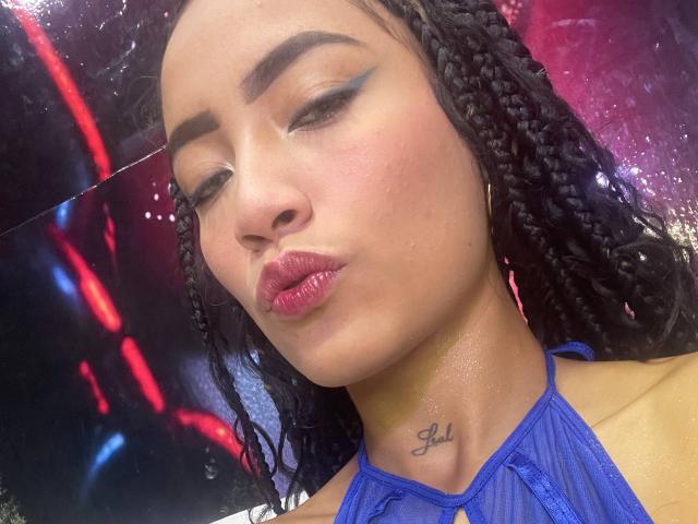 SusyXpleasure tiene el cabello oscuro trenzado, delineador de ojos azul, labios rosados brillantes y un pequeño tatuaje con la palabra "Lola" en el cuello. Lleva un top azul con tirantes..