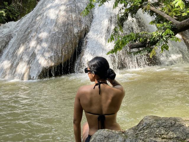 RoxyThunder con i capelli neri raccolti in uno chignon, indossa un bikini nero, seduta su delle rocce, di fronte a una cascata, con gli occhiali da sole, di spalle alla telecamera, con l'acqua che schizza intorno a lei..