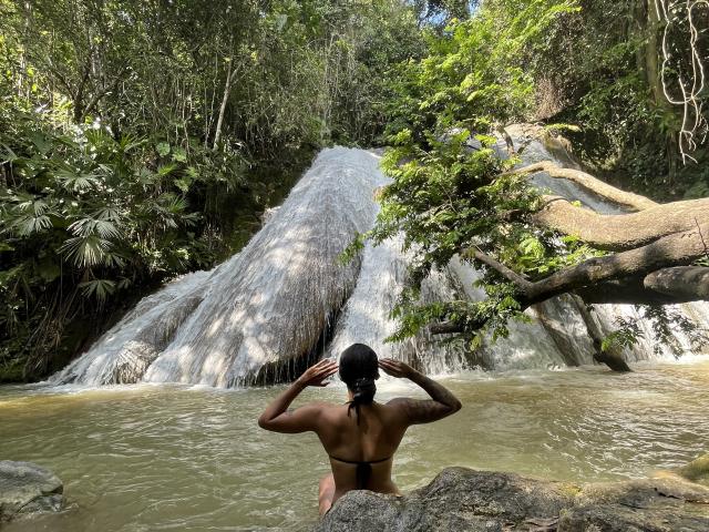 RoxyThunder with dark skin and black hair in a bun, wearing a bikini, stands in a waterfall's pool, hands on head, looking up at the cascading water.