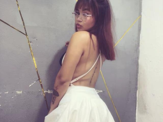 EstefyChaud, avec une partie de sa poitrine visible, regardant par-dessus son épaule, portant des lunettes, une jupe blanche et un haut à bretelles, exposant son dos et ses fesses, avec un tatouage sur son bras droit..