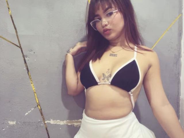 EstefyChaud con gli occhiali, reggiseno nero, gonna bianca, piccolo tatuaggio a forma di farfalla sul petto, piercing al naso, capelli castani e pelle chiara, in piedi contro un muro grigio con una corda gialla..