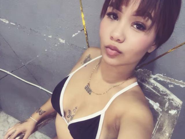 EstefyChaud hat kurze, braune Haare, trägt einen schwarzen und weißen Bikini-Oberteil, auf dem kleine Tattoos auf ihrer Brust und ihren Armen zu sehen sind, und sie trägt eine goldene Halskette und hat einen schmollenden Gesichtsausdruck..