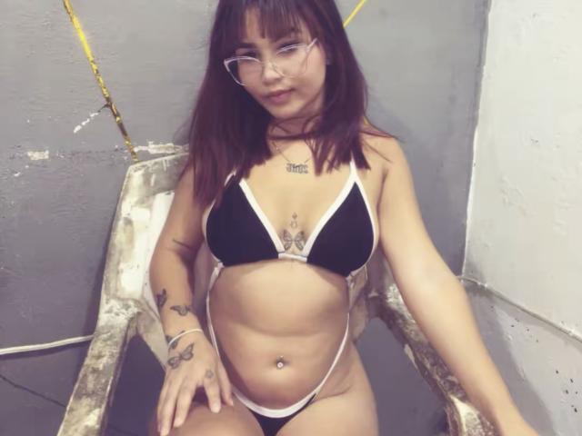 EstefyChaud, con cabello castaño y gafas, vestida con un bikini blanco y negro, sentada en una silla desgastada, mostrando tatuajes y un piercing en el ombligo, mirando ligeramente hacia abajo..