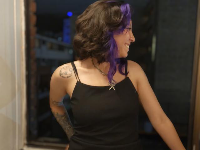 DarckVelvet with purple and black hair, black tank top, tattoos on arm and shoulder, smiling, wearing star necklace, standing near door, dark background, slightly wavy hair.
