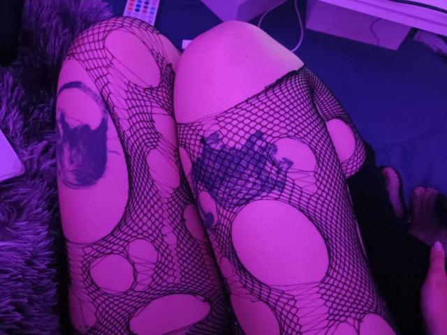 DarckVelvet's legs in black fishnet stockings with large circular holes, revealing tattoos on both thighs. Purple lighting. Her ass is partially visible, showing her fishnet-covered skin.