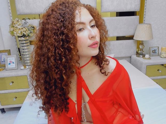 IrisDalton with curly red hair, wearing a sheer red top, showing her small tits and cleavage, sitting on a bed, looking slightly to the side, in a bright room.