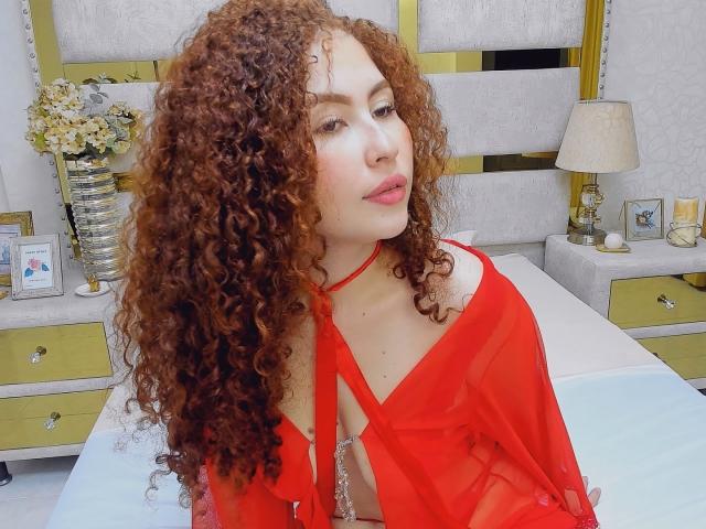 IrisDalton with long curly red hair, wearing a sheer red top, exposing her tits, sitting on a bed, looking slightly to the side with pink lips.