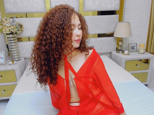 IrisDalton with curly red hair, wearing a red sheer top that reveals her tits and a tattoo on her side, sitting on a bed, eyes closed, looking sexy.