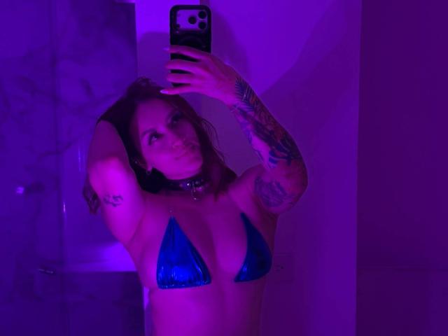 MaraSantoro with blue triangle bikini, arms raised, tattoos visible, purple lighting, taking selfie, wearing choker, holding phone, looking at mirror, showing tits, in bathroom.
