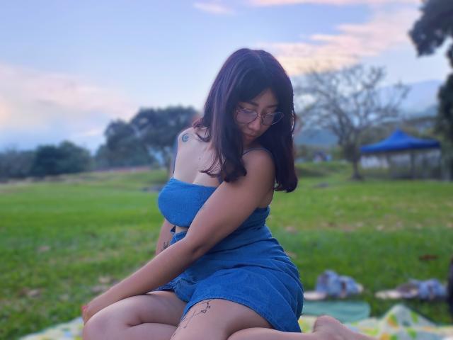 Genesiss with dark brown hair, glasses, and a blue tube top and shorts, sitting on grass, showing her tattooed arm and small tits, with a shy smile.
