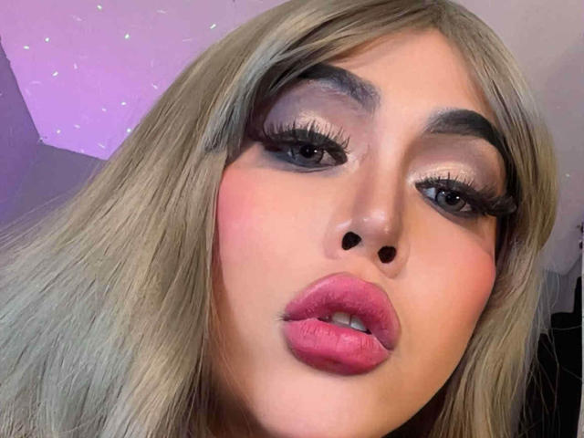 Le visage inoubliable de Maria, avec ses cheveux blonds, son rouge à lèvres rose et son eye-liner prononcé, faisant la moue avec ses lèvres pleines et brillantes, et ses longs cils épais et artificiels. Elle regarde directement l'appareil photo..