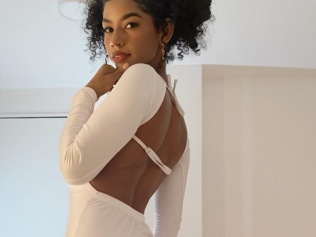 Litalexa with dark curly hair, brown skin, wearing a white backless dress, looking over her shoulder, fingers near her mouth, showcasing her bare back and white straps.