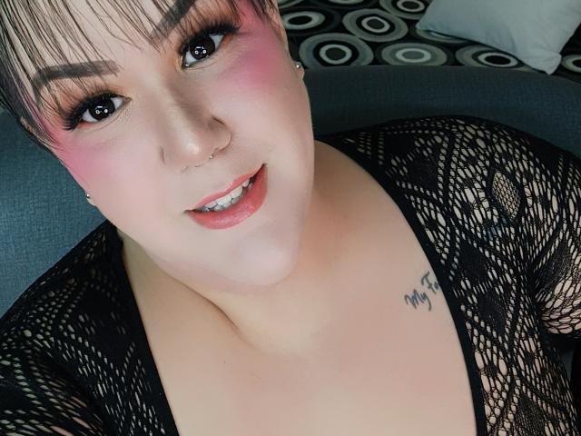 GiiannaMorrys mit rosigem Rouge, schwarzem Netzoberteil, Nasenring und Tattoo auf der Brust, lächelnd mit sichtbaren Zähnen. Kurzes, braunes Haar, dunkler Eyeliner und rosiger Lippenstift..