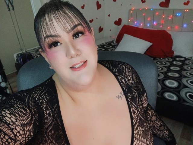 GiiannaMorrys con top in rete nera, fard rosa, eyeliner e piercing al naso, sorride alla fotocamera, mostrando un piccolo tatuaggio sul lato destro del petto, seduta su una sedia grigia..