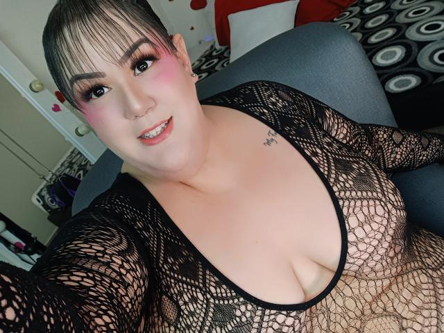 GiiannaMorrys mit rosigem Rouge, schwarzem Netzoberteil, das große Brüste zeigt, Tattoo an der rechten Schulter, lächelnd, dunkles Haar zu einem Dutt gebunden, sitzend auf einem grauen Sofa, Nasenring..