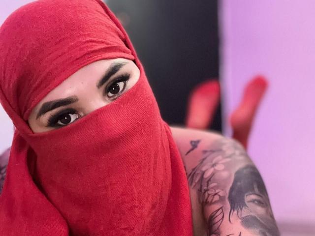 MalakCote, avec un foulard rouge et un masque, montrant son bras tatoué et ses sourcils épais et foncés, allongée, les yeux fixés vers l'avant. Des chaussures rouges en arrière-plan, un mur violet..