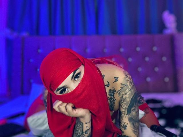 MalakCote con un pañuelo rojo en la cabeza, tatuajes que cubren los brazos y el pecho, mirando a la cámara, acostada en la cama, con iluminación púrpura y azul, vistiendo lencería roja, con la mano debajo del mentón..