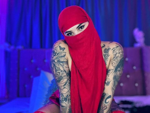 MalakCote, avec ses tatouages, portant un masque rouge et une écharpe rouge assortie, est assise dans une pièce éclairée en bleu, mettant en valeur ses bras et ses jambes tatoués, avec un regard sexy et mystérieux..