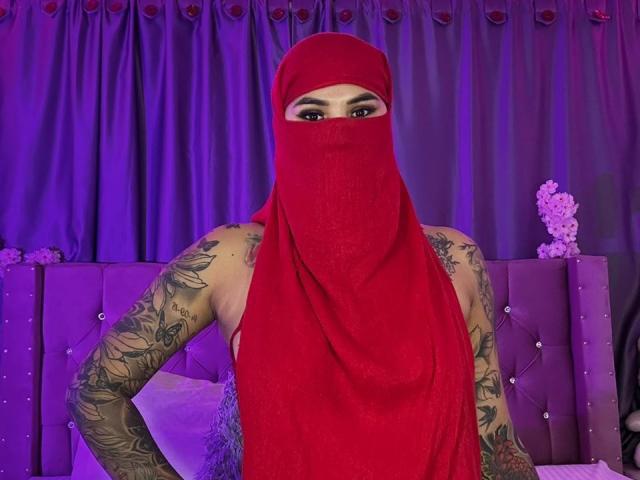 MalakCote, avec des tatouages sur les bras, portant un masque de ninja rouge et un tissu rouge recouvrant le visage et le corps, se tient devant une tête de lit tuftée avec une teinte violette..