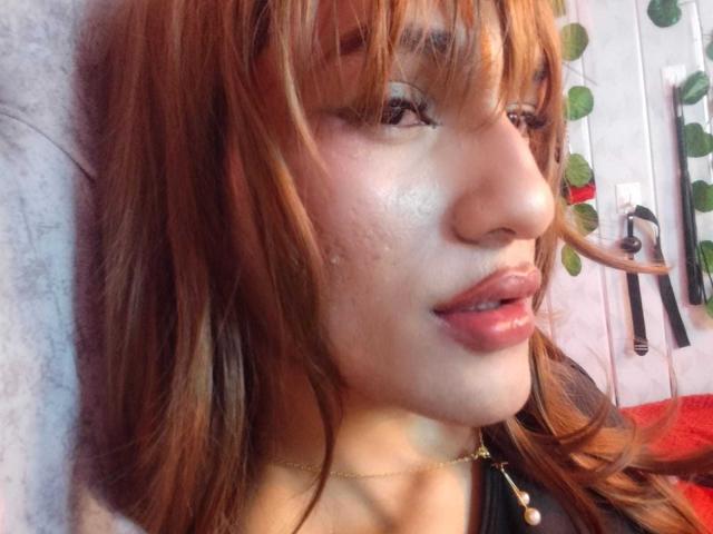 PamelaXFox with red hair, shiny skin, and full pink lips, wearing a black top, has a gold necklace, and a slight smile showing her teeth.