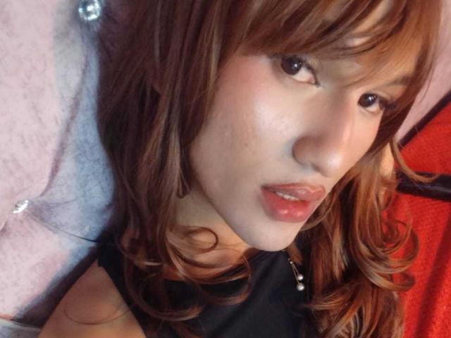 PamelaXFox a les cheveux bruns clairs et ondulés, un joli visage et des lèvres roses brillantes. Elle porte un haut noir avec un collier de perles, et ses fesses sont partiellement visibles..
