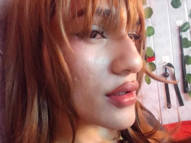 O rosto de PamelaXFox, com cabelo castanho claro e ondulado com franja, lábios rosados, algumas sardas e um sorriso discreto, com um nariz visível e um colar de ouro..