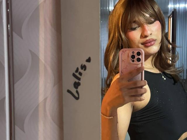 PamelaXFox con cabello castaño ondulado, top negro sin mangas, sosteniendo un teléfono rosa con cámara blanca, tomando una selfie, ligeramente sonrojada, usando una pulsera, lápiz labial rosa, trasero pequeño..