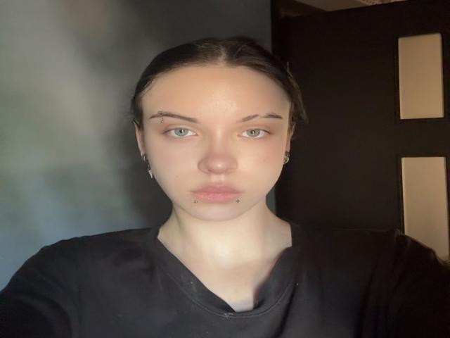 RosendaLazcano has light skin, green eyes, and a serious expression. She's wearing a black shirt with small earrings and has lip and nostril piercings, plus a chin piercing.