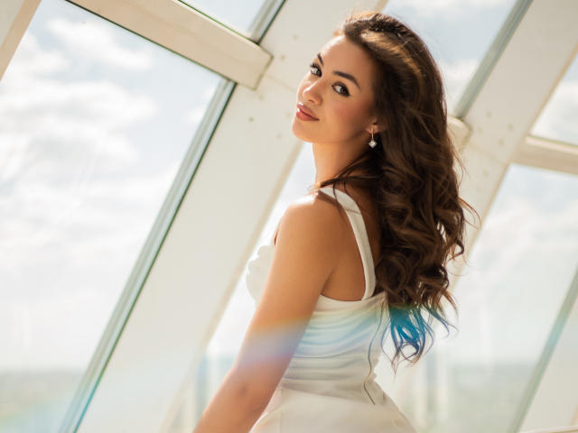 EricaSiin with long wavy brown hair, wearing a white sleeveless dress, looking over her shoulder, smiling, showing her tits, standing by a sunlit window.