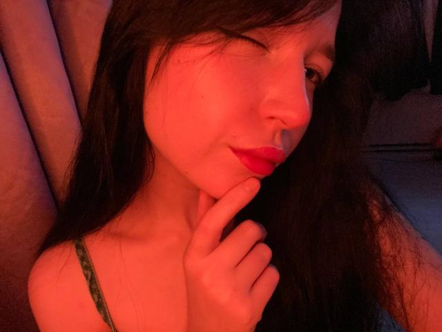 MertieDasinger with long black hair, winking, red lips, green strap, touching chin, soft red light, slightly smiling, looking to the side.
