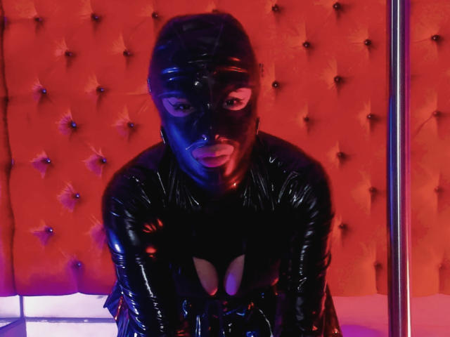 RoxanneHan in black latex mask and outfit, crouching, showing cleavage, in red tufted background, with metal pole on right. Her lips are slightly parted, looking forward.