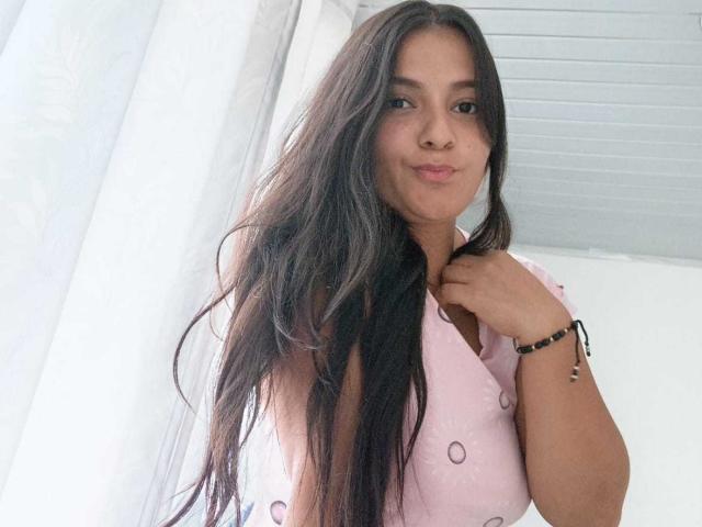 GiaaSmith with long dark hair, pink dotted top, smiling slightly, right hand on chest, black bracelet, standing near white curtains, ceiling visible.