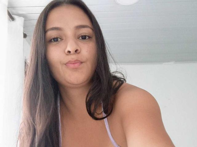 GiaaSmith with long brown hair, light brown skin, small nose, and puffy lips, wearing a purple spaghetti-strap top, looking at the camera with a slight smile.