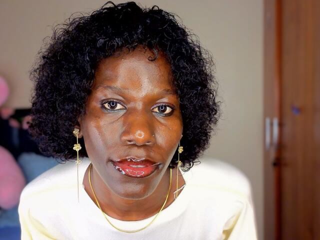 KharlaQueen has dark curly hair, wearing gold earrings and a white shirt, with a slight smile and red lipstick, looking directly at the camera.