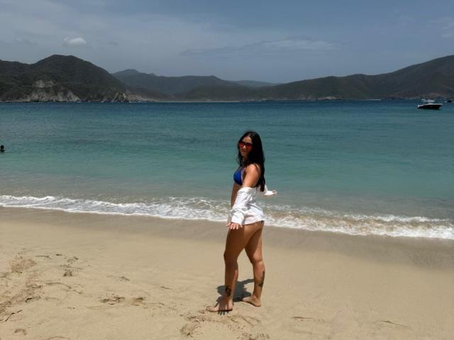 JuliaGarciax mit langen, schwarzen Haaren, die rote Sonnenbrillen, einen blauen Bikini-Oberteil und ein weißes Hemd trägt, steht auf einem sandigen Strand und zeigt ihr Gesäß, im Hintergrund sind ein grün-blauer Ozean und Berge zu sehen..